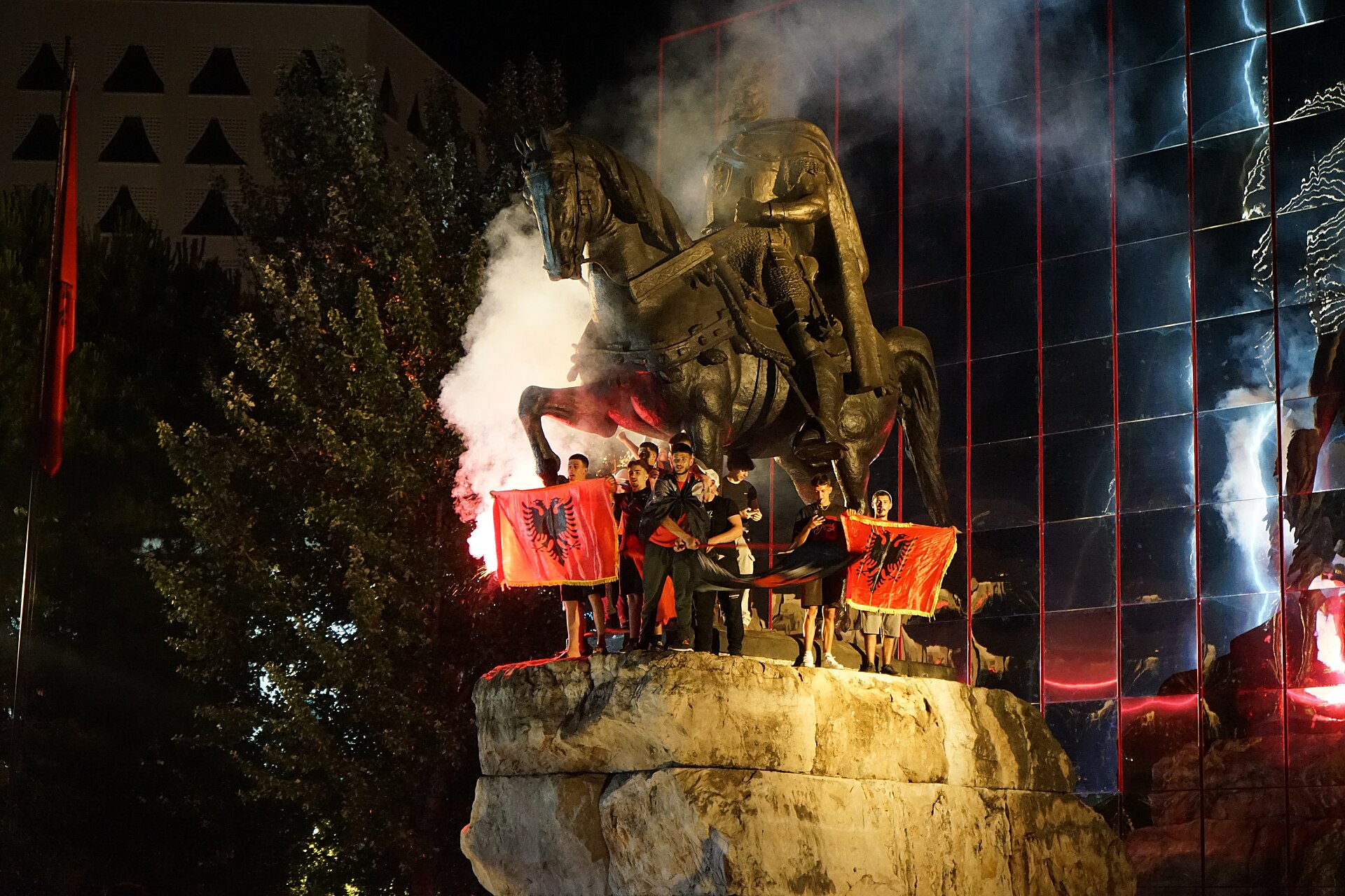 Protesters holding Albanian flags stand on the Skanderbeg monument in Tirana at night amid flare smoke — Albania protests file photograph