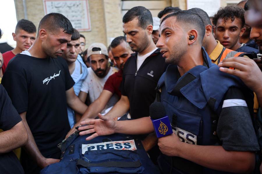 A Gaza reporter in a blue press vest speaks beside a damaged PRESS flak jacket after a journalist was killed