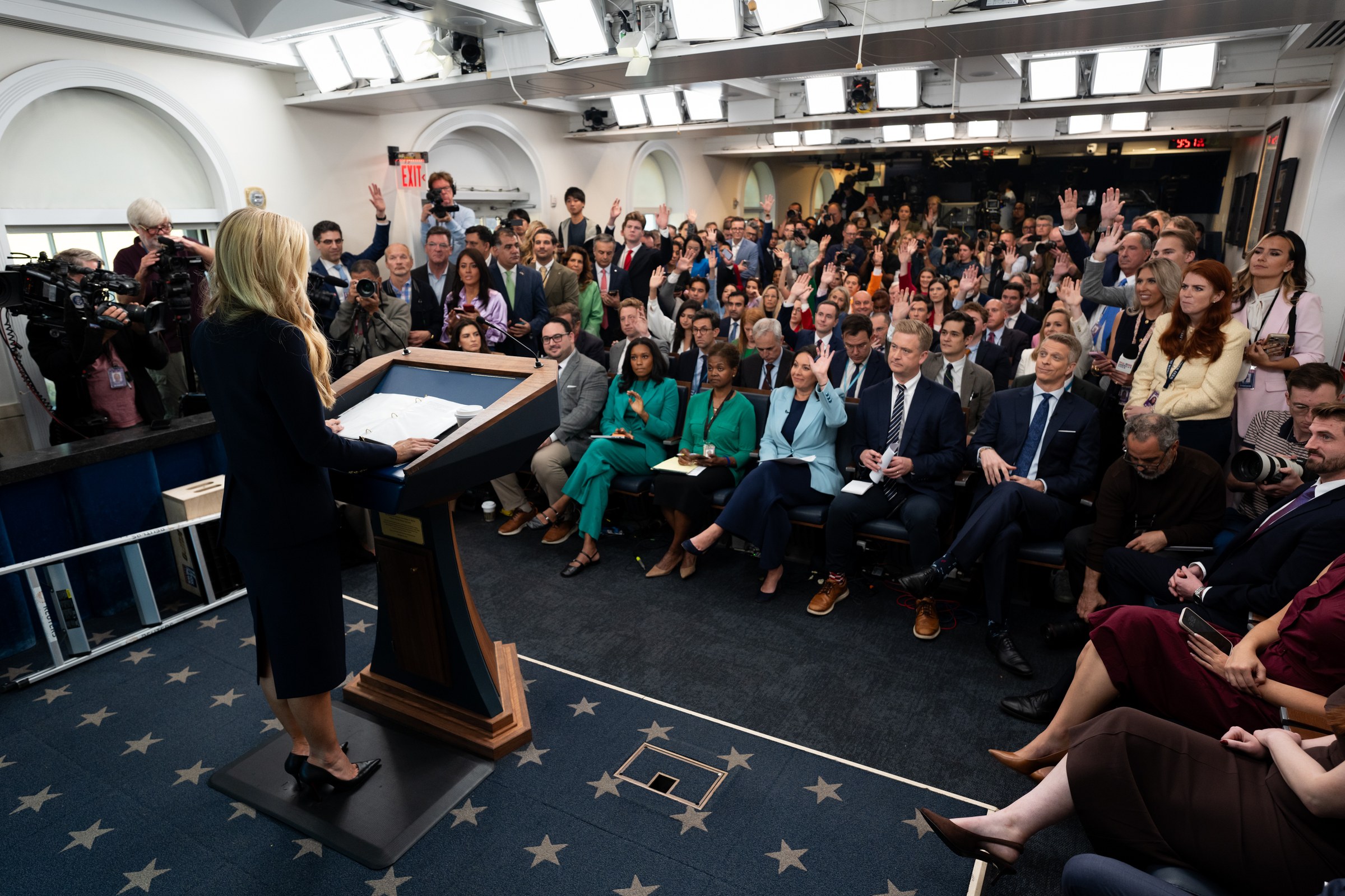 Katie Zacharia at the podium facing rows of reporters with raised hands during her first White House briefing