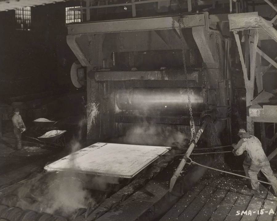 Steelworkers guide a hot slab through rolling equipment at Gary Works, illustrating the scale of the planned Mesabi Metallics Iowa steel plant