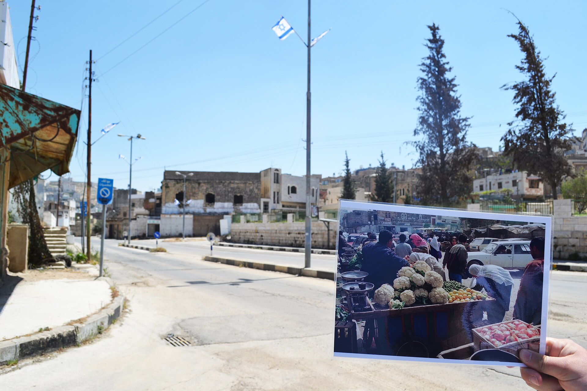 Palestinians displaced in Hebron — Shuhada Street in Hebron's Old City standing empty under military closure, a held-up photograph showing the same corner as a busy market in 1999