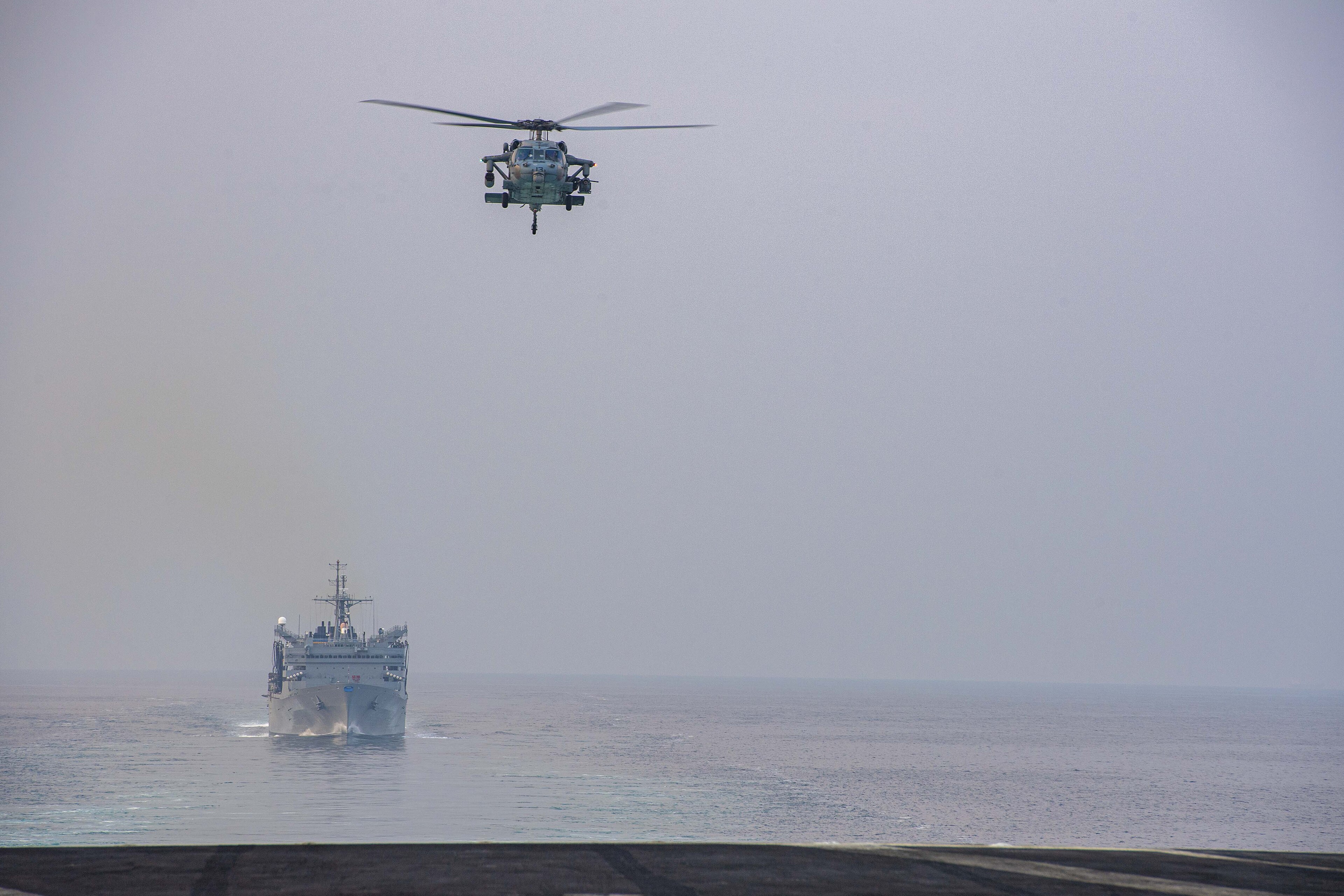 A U.S. Navy MH-60S Sea Hawk helicopter flies above a fast combat support ship as the Eisenhower Carrier Strike Group transits the Strait of Hormuz