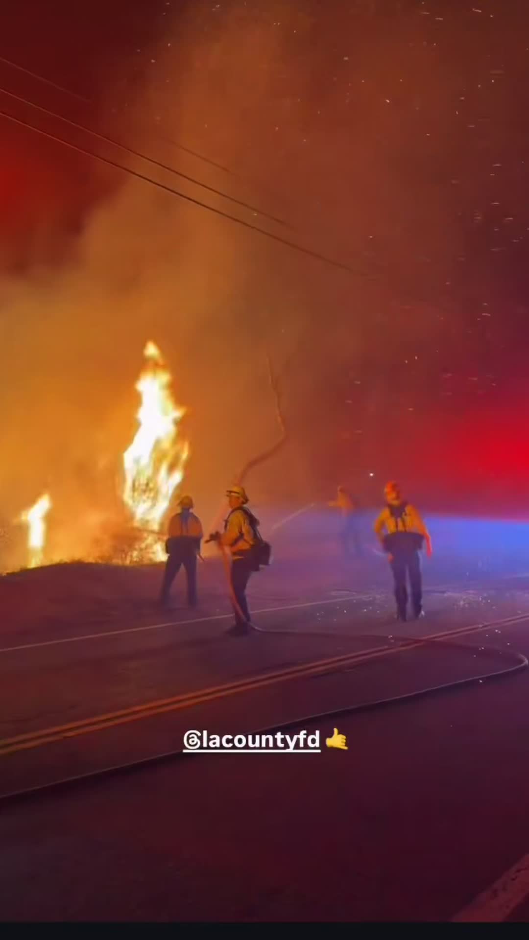 Los Angeles County firefighters work a hose line on a canyon road at night beside a wall of flame from the Bryson Fire