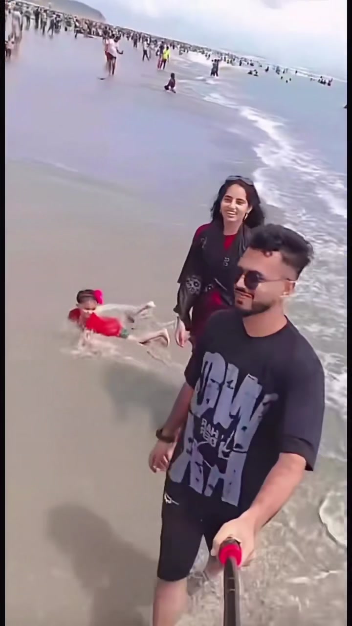 The toddler in a red top in the shallows as a wave washes over her, metres behind her parents' camera