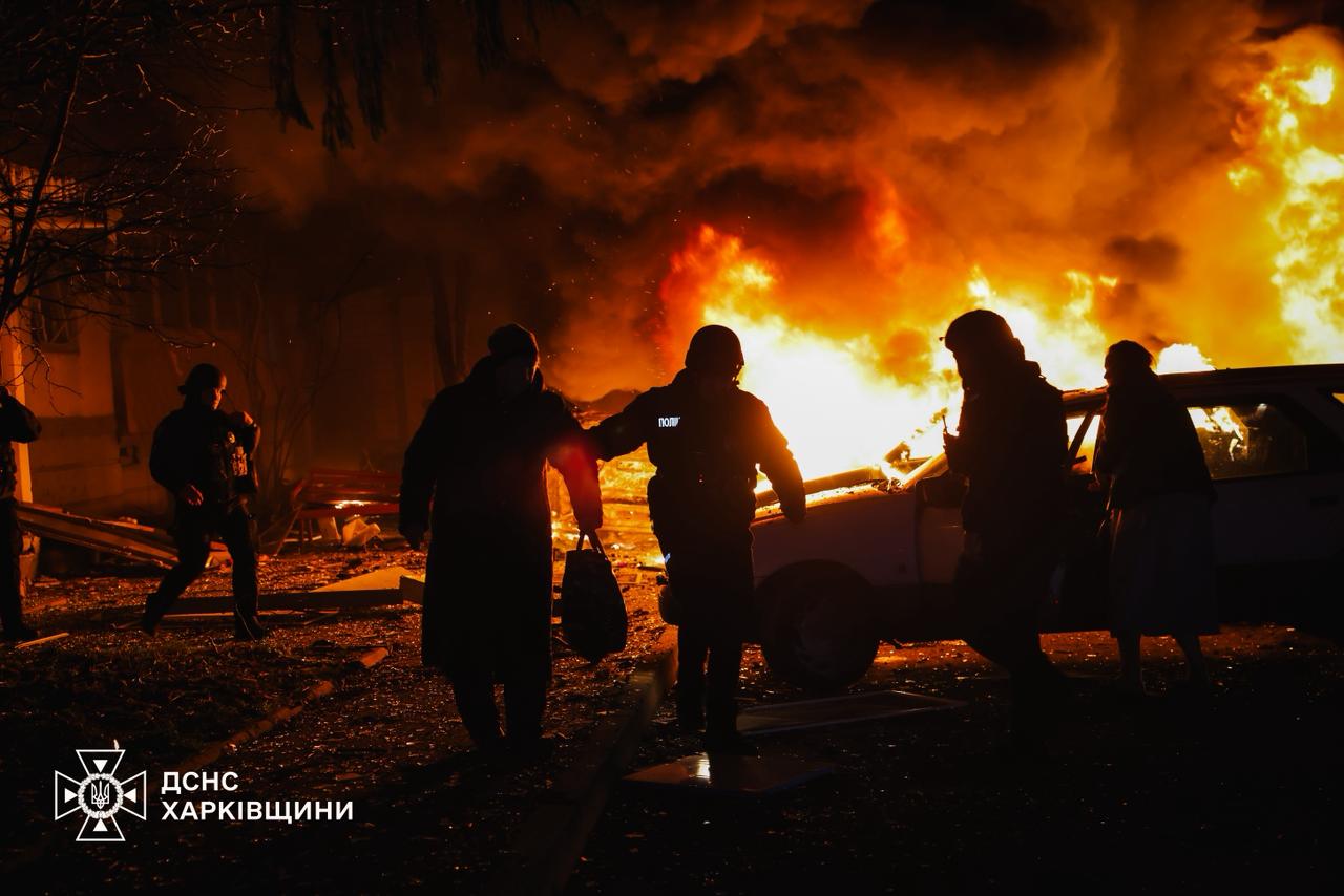 Silhouetted residents and a police officer walk past a burning car in Kharkiv after a drone strike at night — State Emergency Service file photo from a November 2025 attack on the city