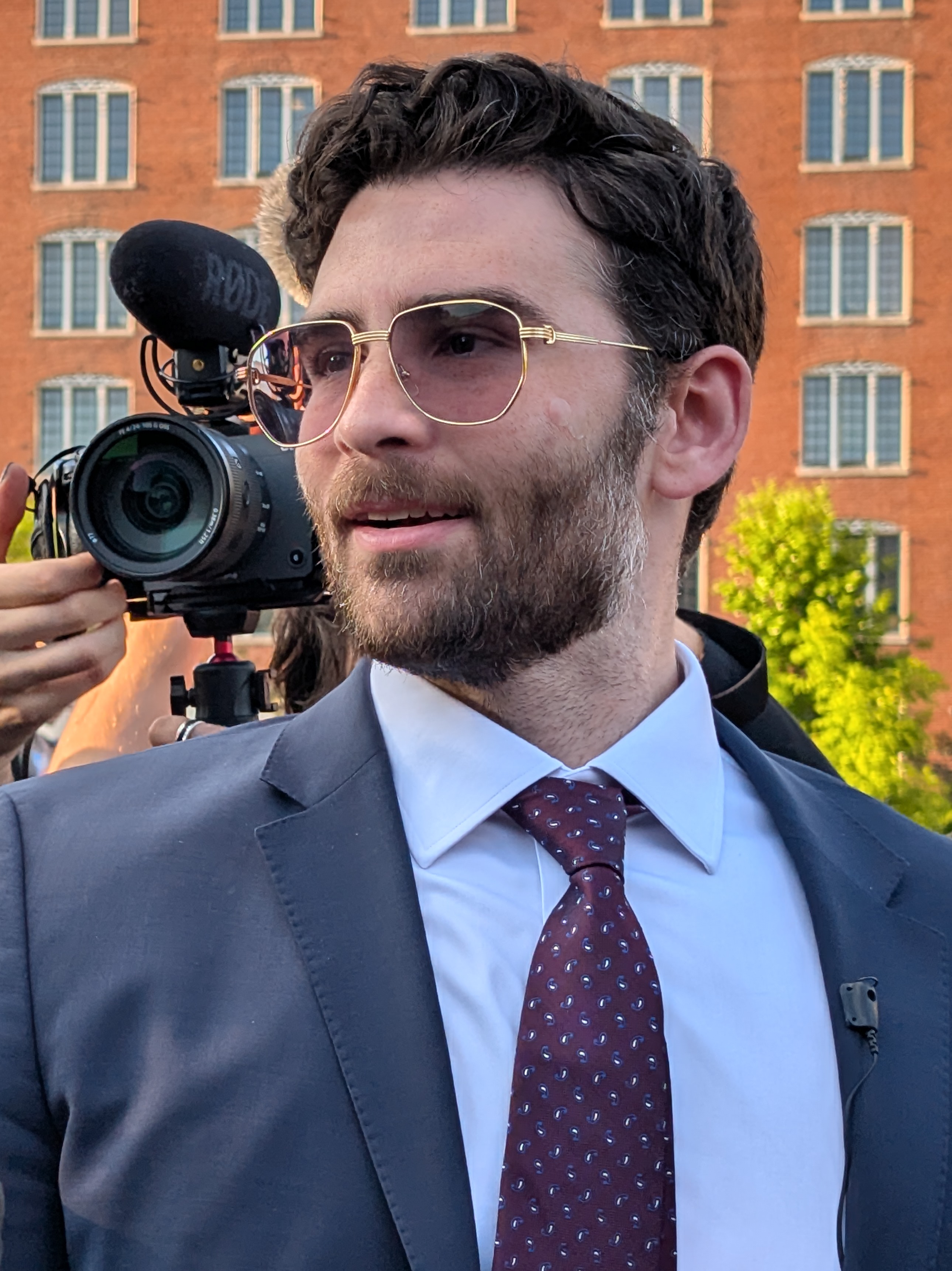 Hasan Piker sues Scott Jennings — Hasan Piker in sunglasses, a suit and a maroon tie at a rally in St. Louis in 2026, a camera operator behind him