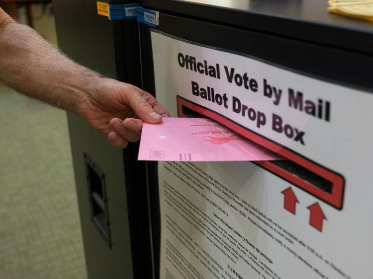 A voter&rsquo;s hand placing a pink vote-by-mail ballot envelope into an official California ballot drop box