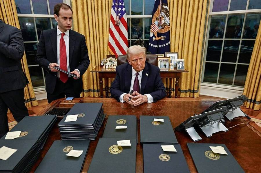 President Donald Trump seated at the Resolute Desk in the Oval Office while signing executive orders