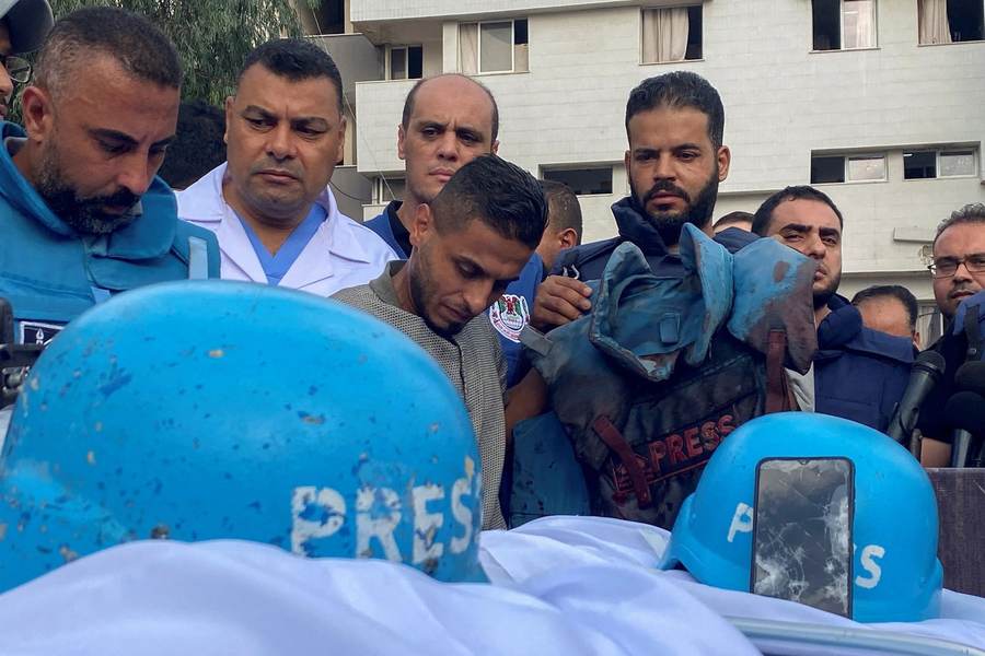Journalists stand behind damaged blue PRESS helmets and a bloodstained flak jacket at a Gaza hospital memorial