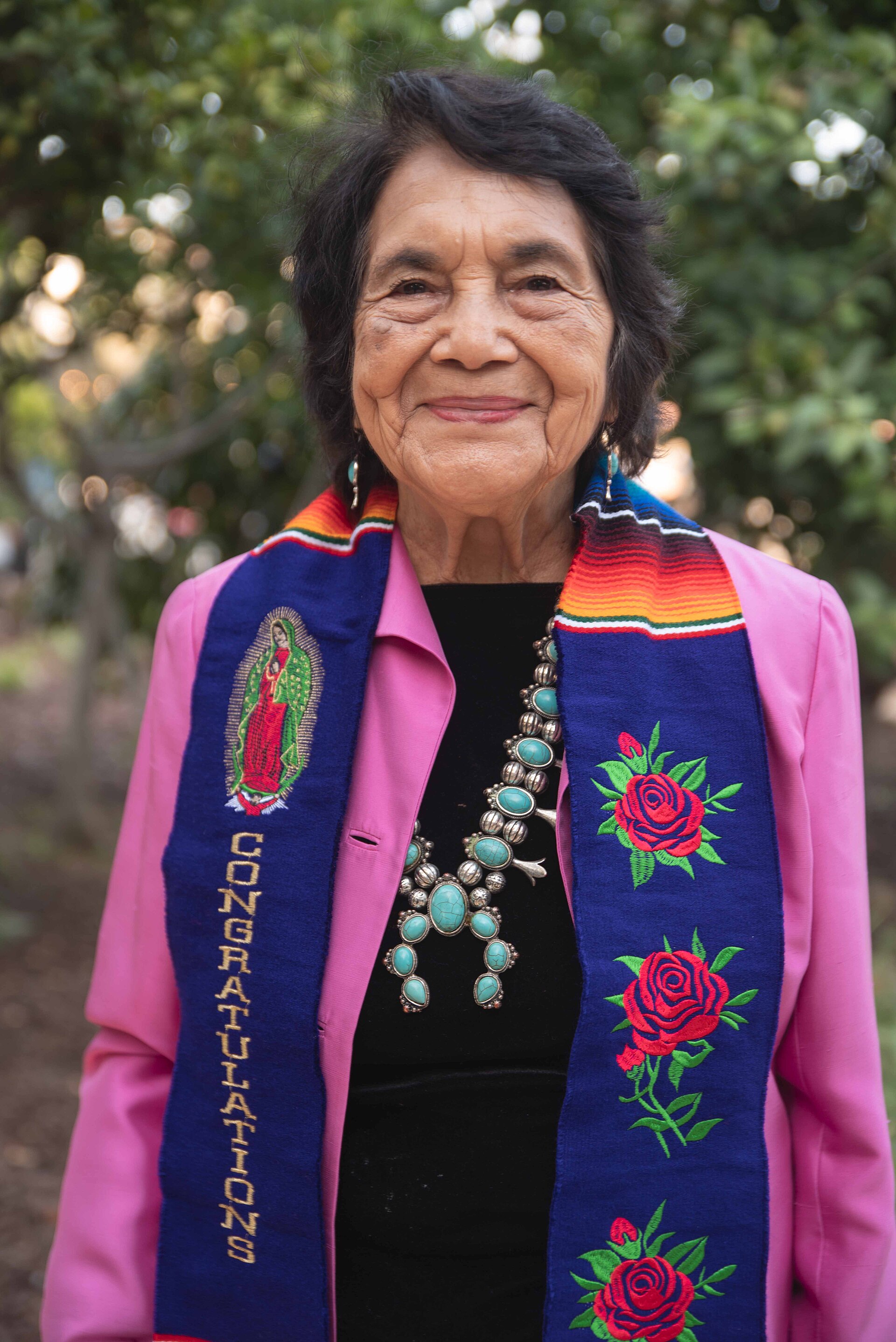 Dolores Huerta in 2023, smiling outdoors in a pink jacket and an embroidered blue 'Congratulations' stole as she celebrates her honorary doctorate from USC