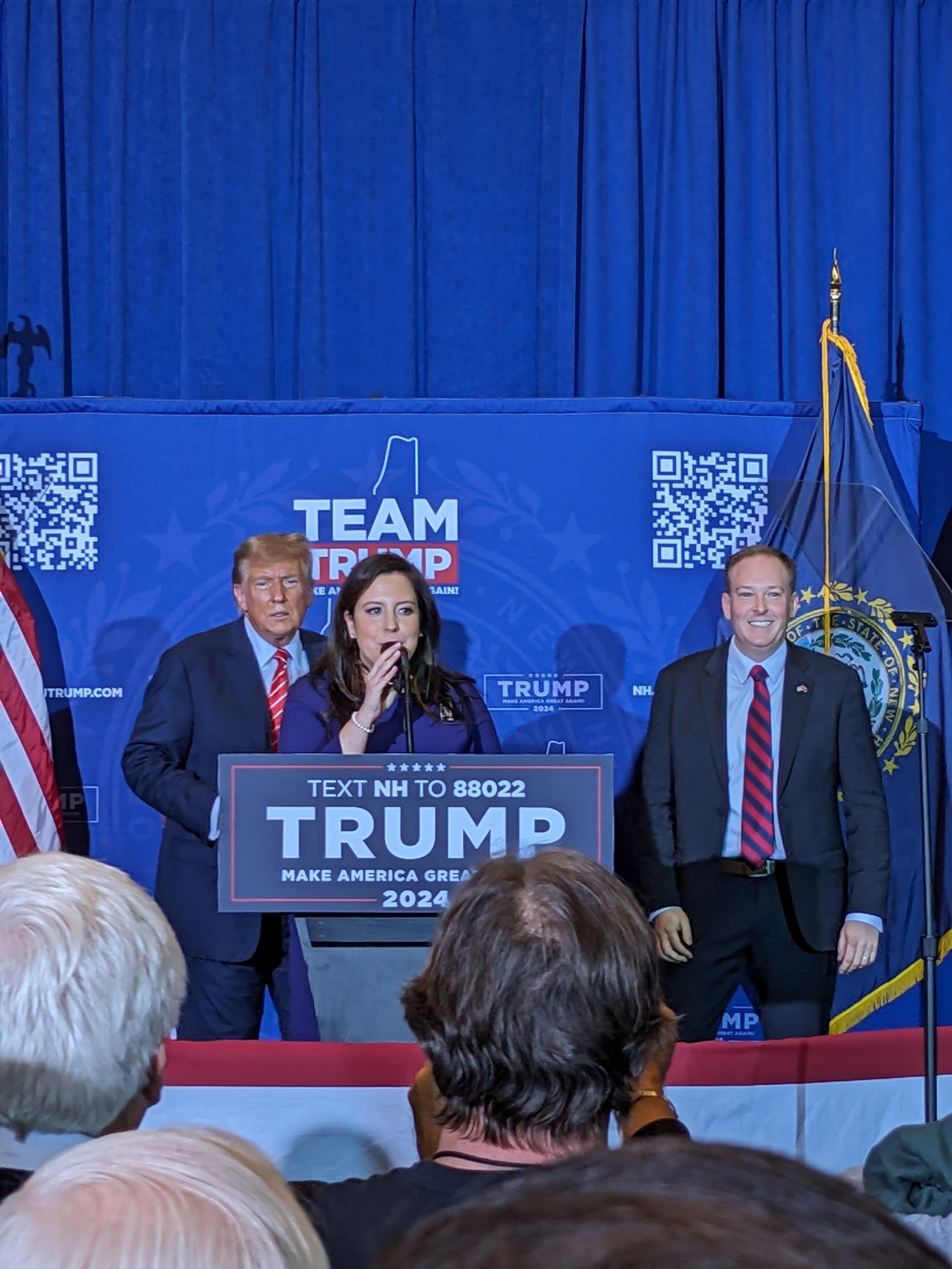 Donald Trump standing behind Representative Elise Stefanik at the podium of a campaign rally in Concord, New Hampshire, with supporters in the foreground