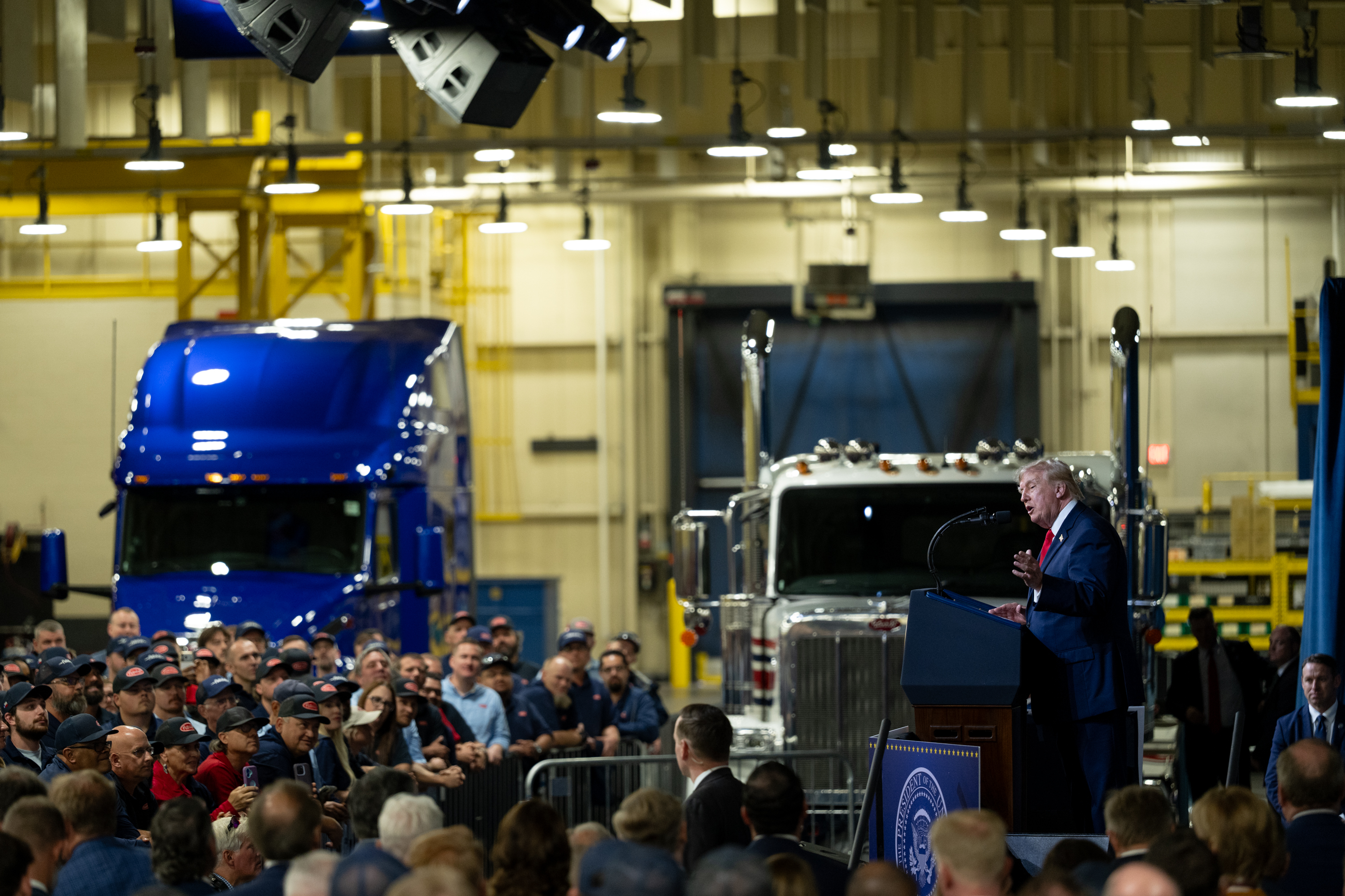 President Donald Trump speaking at a podium to an audience inside the Peterbilt Motors plant in Denton, Texas
