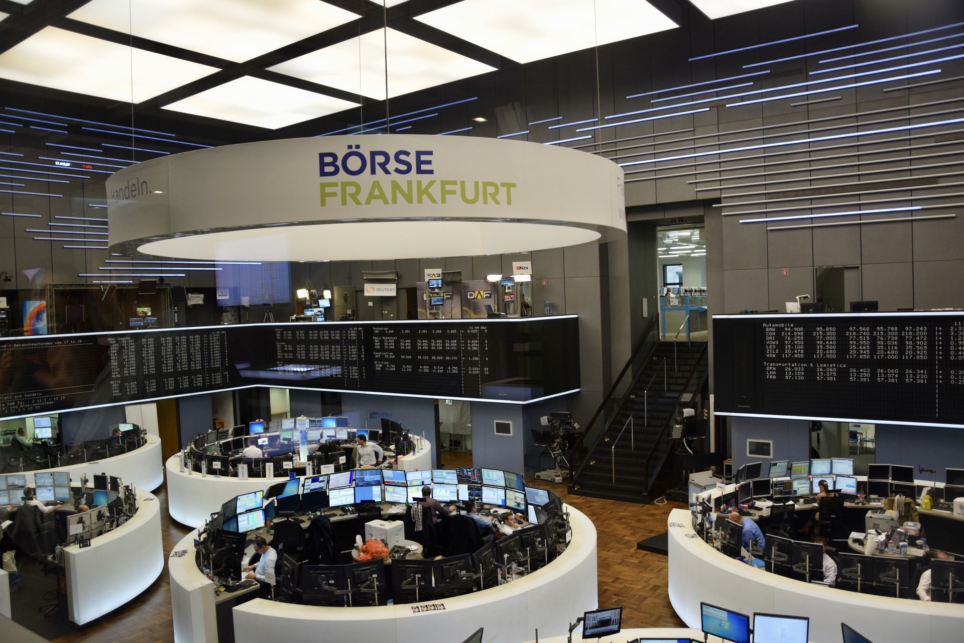 A stock exchange trading floor with rows of desks and monitors beneath a large electronic index board showing market data