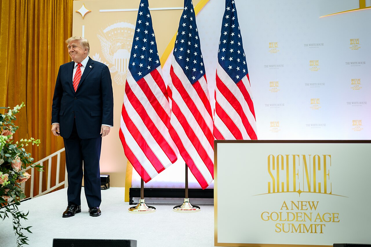 President Donald Trump standing on stage beside US flags at the New Golden Age summit in Washington on October 8, 2026